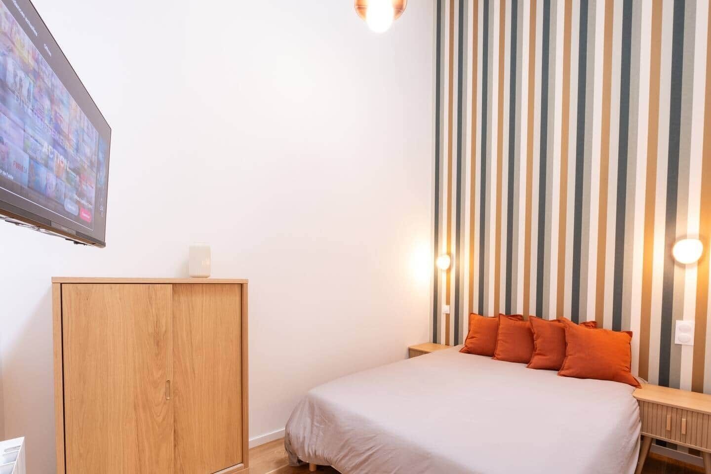 Une chambre d’hôtel avec un lit, une armoire en bois, un téléviseur fixé au mur et un papier peint à rayures.