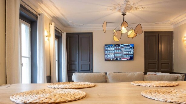 Un comedor con mesa de madera, individuales tejidos, una araña y un televisor en la pared.