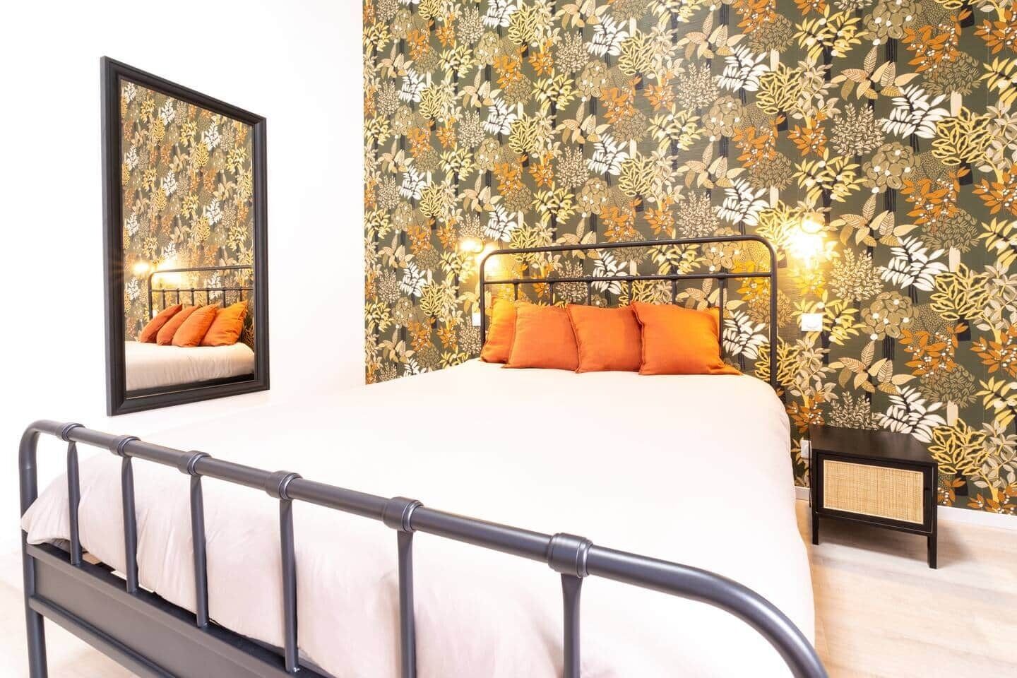 A bedroom with a bed, a nightstand, a mirror, and a floral wallpaper.