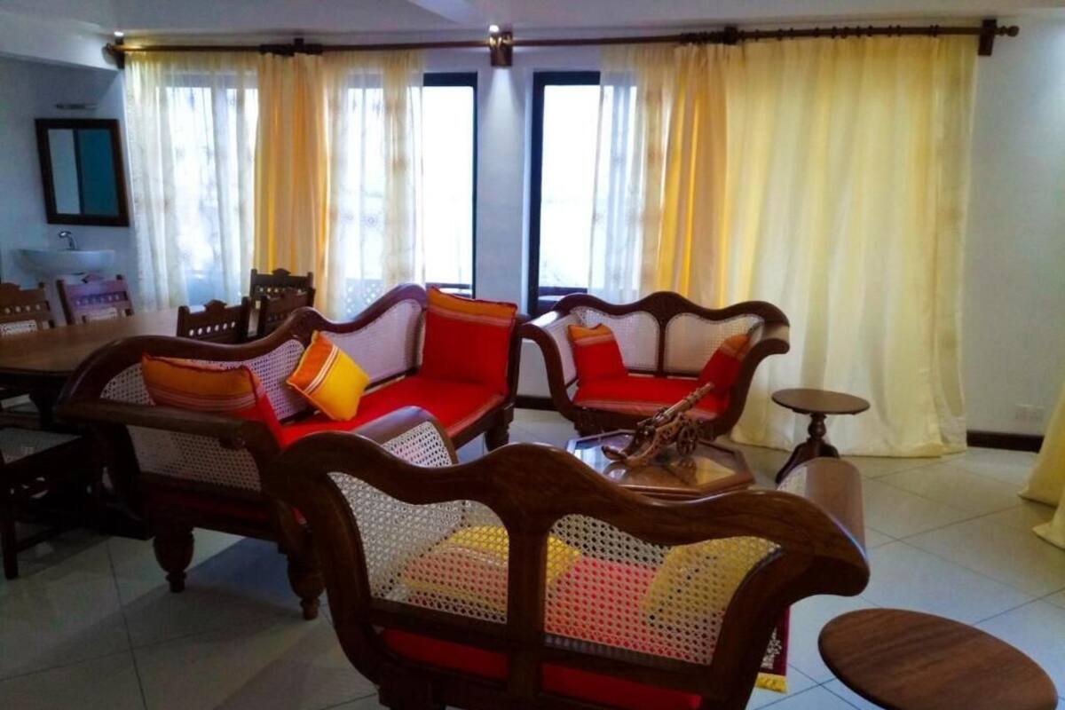 A living room with wooden sofas and a round table, large windows with yellow curtains, and a dining area with chairs and a table.