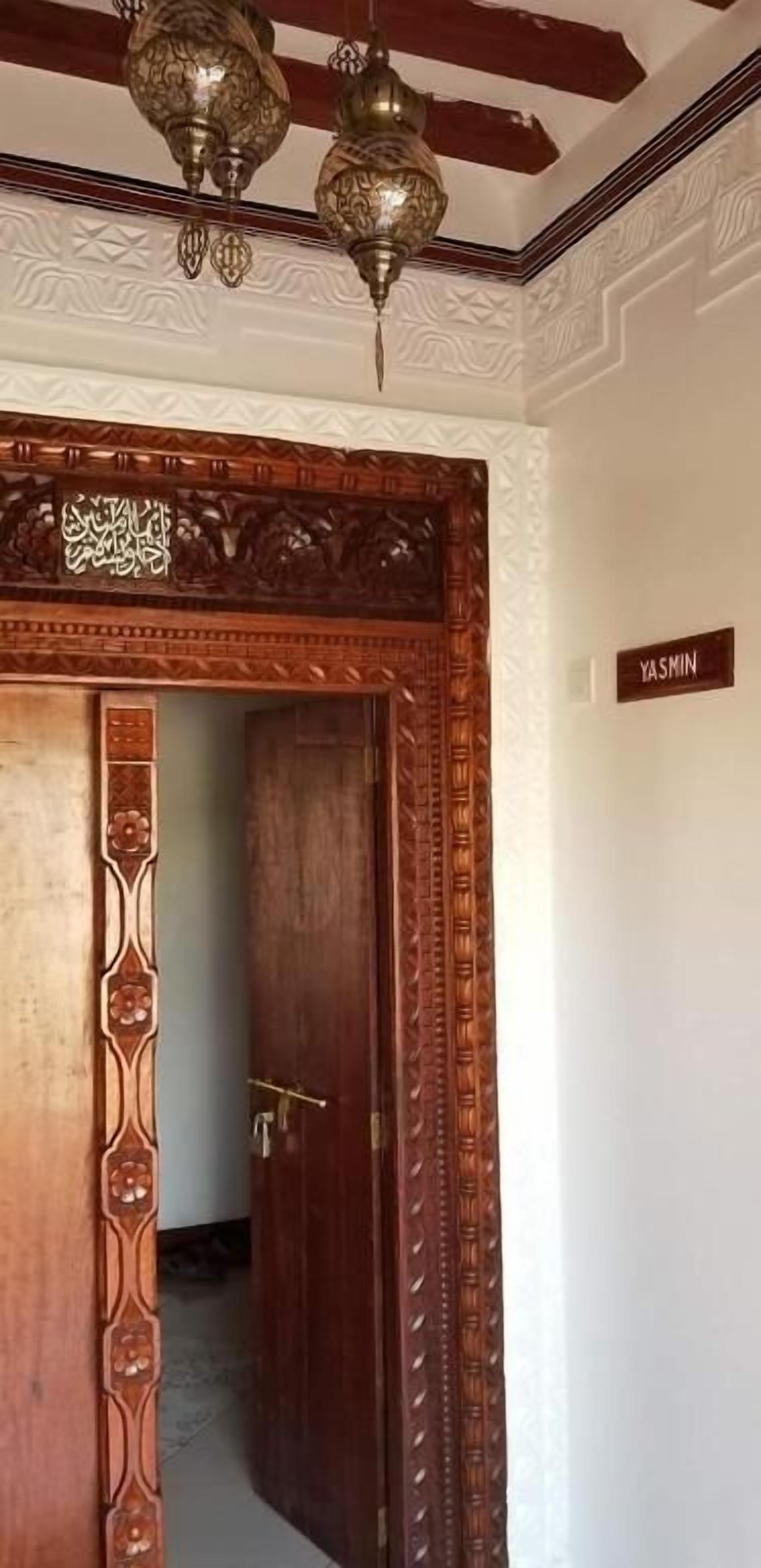 Eine Holztür mit aufwendigen Schnitzereien und darüber eine Messing-Chandelier. An der Wand ist ein Schild mit dem Schriftzug "YASMIN" zu sehen.