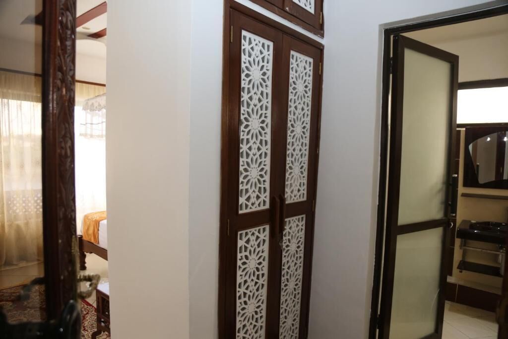 Una habitación con un armario de madera decorado con diseños tallados, una cama con cubrecama blanco y un baño visible con espejo y lavamanos.