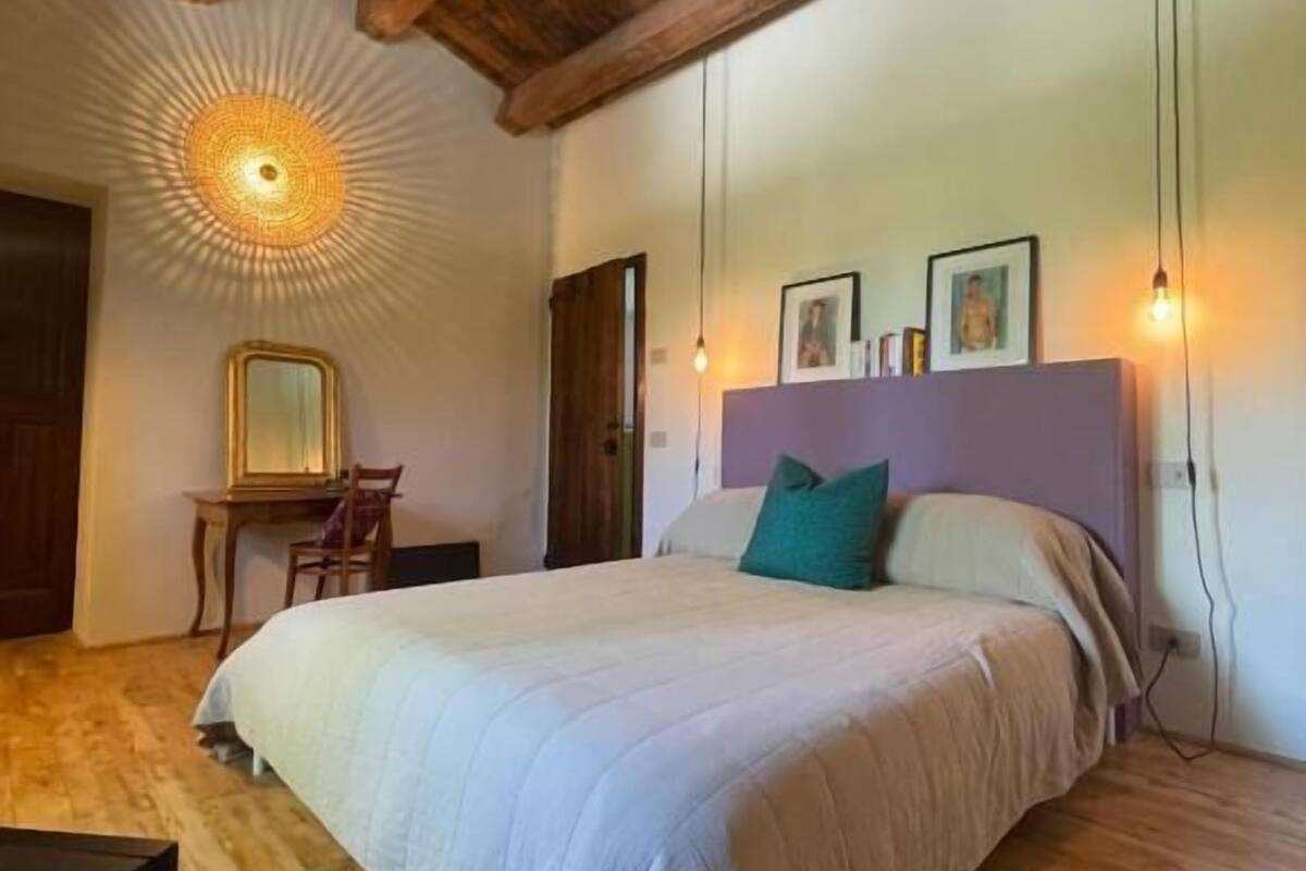 A bedroom with a wooden ceiling, a bed with a white and teal bedspread, a desk with a mirror, and a wall-mounted light fixture.
