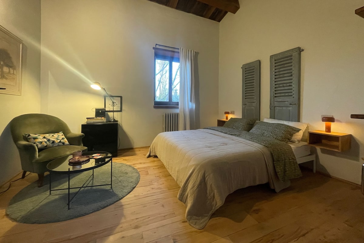 A bedroom with a bed, a chair, a small table, a window, and a radiator.
