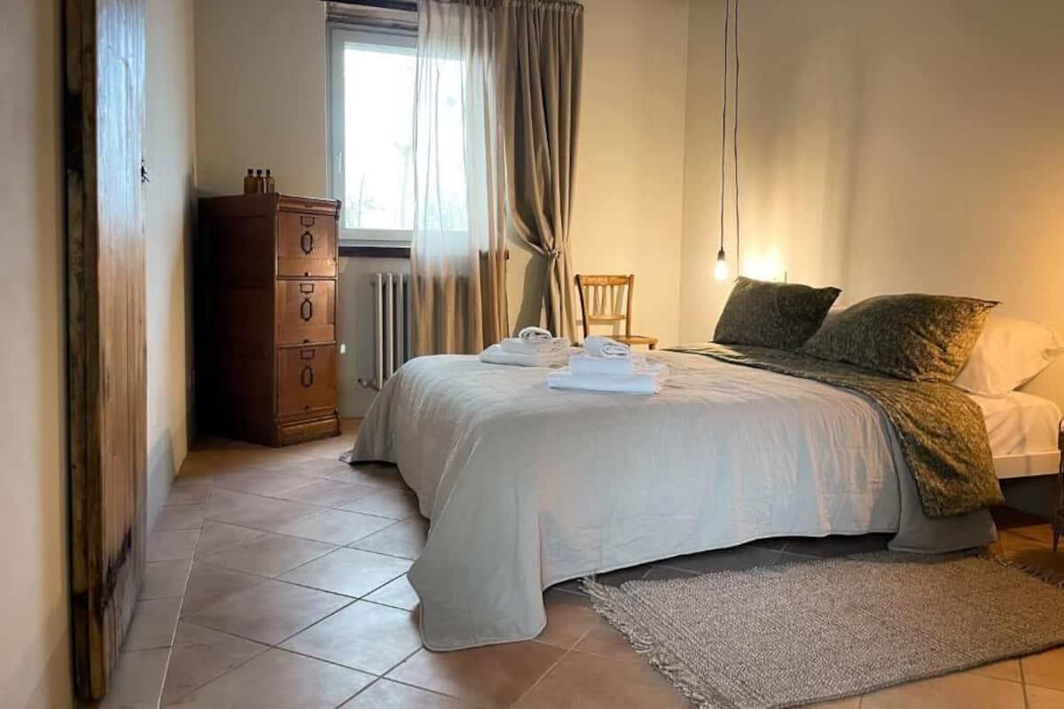 A bedroom with a bed, a wooden dresser, a chair, and a window with curtains.