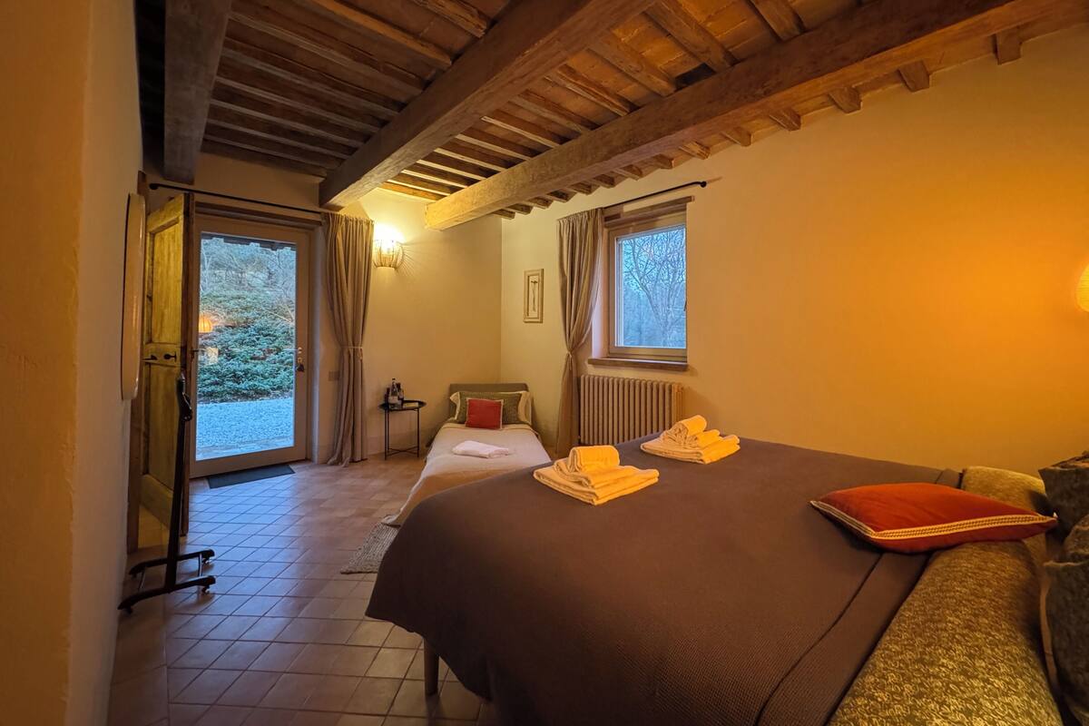A cozy room with a wooden ceiling, a bed with a brown bedspread, two windows with curtains, and a small table with a bottle and glasses.