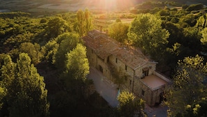 Un bâtiment en pierre, doté d’un toit en tuiles, est entouré d’une végétation luxuriante et s’élève sur un arrière-plan de collines.
