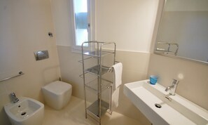 Ein Badezimmer mit einem weißen Waschbecken, einem Bidet, einem Metallregal und einem Fenster.