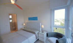 Ein Schlafzimmer mit einem Bett, Nachttischen, einem Sessel, einem Gemälde und einem Fenster mit Aussicht.