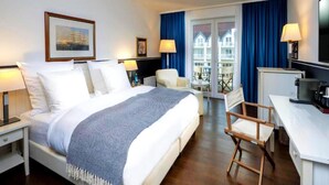 A hotel room with a bed, a desk, a chair, a TV, and a balcony with blue curtains. - Sunday Hafendorf Double Room With Lake-view (Rheinsberg)