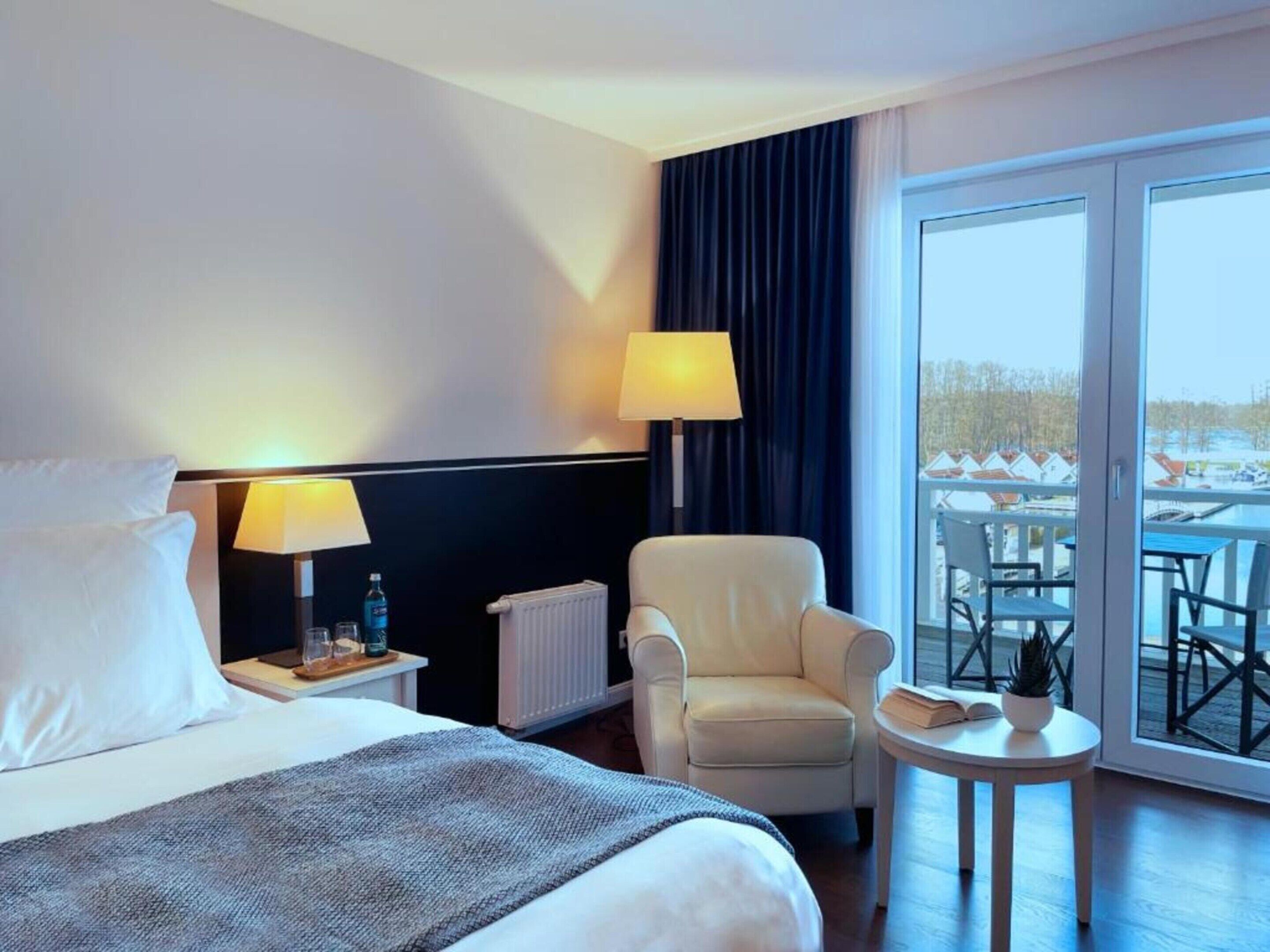 Ett hotellrum med en säng, två sänglampor, en stol, ett litet bord med en växt och utsikt över en balkong med utomhusmöbler.