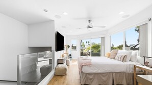 Ein modernes Schlafzimmer mit einem großen Bett, einem Fernseher und einem Balkon mit Blick auf Palmen.