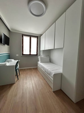 Een kleine kamer met een bed, een bureau en een aan de muur bevestigde televisie.