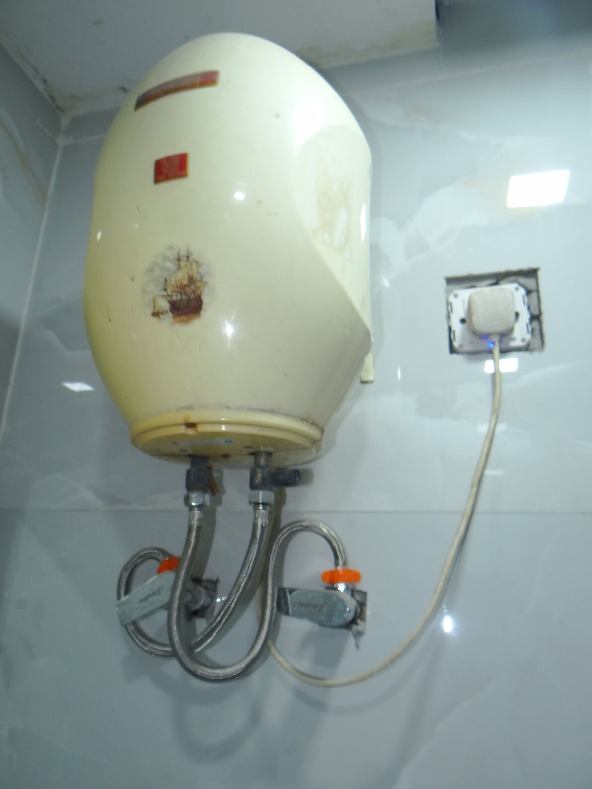 Un chauffe-eau fixé sur un mur recouvert de carreaux, avec des tuyaux apparents et une vanne de contrôle.