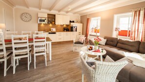 Uma sala de estar espaçosa com um sofá marrom, cadeiras de vime branco, uma mesa de jantar com cadeiras, uma área de cozinha com armários brancos e um relógio na parede.