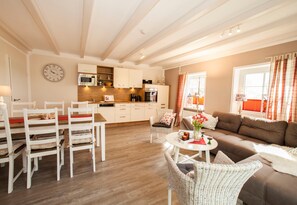Un salon spacieux avec un canapé marron, des fauteuils en osier blancs, une table à manger avec des chaises, un coin cuisine avec des meubles blancs et une horloge au mur.