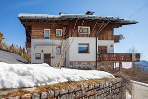 A two-story wooden chalet with a balcony, stone foundation, and a snowy landscape.