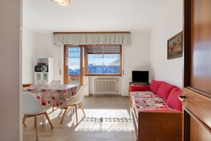 A room with a dining table, chairs, a sofa, a radiator, a television, and a window with a view of mountains.