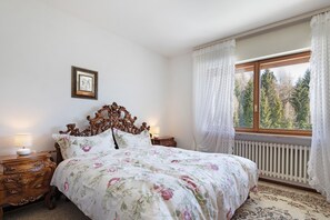 A bedroom with a wooden bed, floral bedding, a nightstand with a lamp, a framed picture on the wall, and a window with curtains.