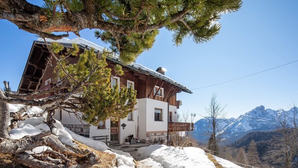 Una cabaña de madera en la montaña con un camino de piedra, rodeada de nieve y pinos, con vistas a una cordillera.