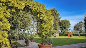 Een park met een pad van baksteen, grote plantenpotten, een houten poppenhuis en een speeltuin met een glijbaan.