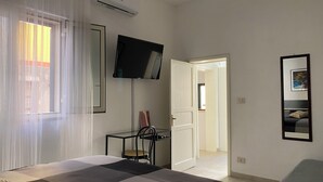Un dormitorio con una cama, un televisor montado en la pared, una silla y una puerta que da a otra habitación.