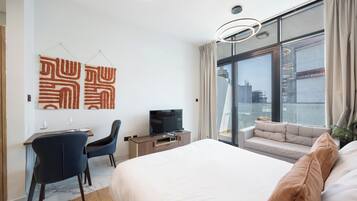 Et moderne hotelværelse med en stor seng, et skrivebord med stol, et fjernsyn og udsigt over byen gennem store vinduer.