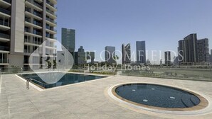 A modern residential building with a rooftop pool area, featuring a large rectangular pool and a circular hot tub, set against a city skyline. - Elegance Stu Al Maryah Vista (Abu Dhabi)