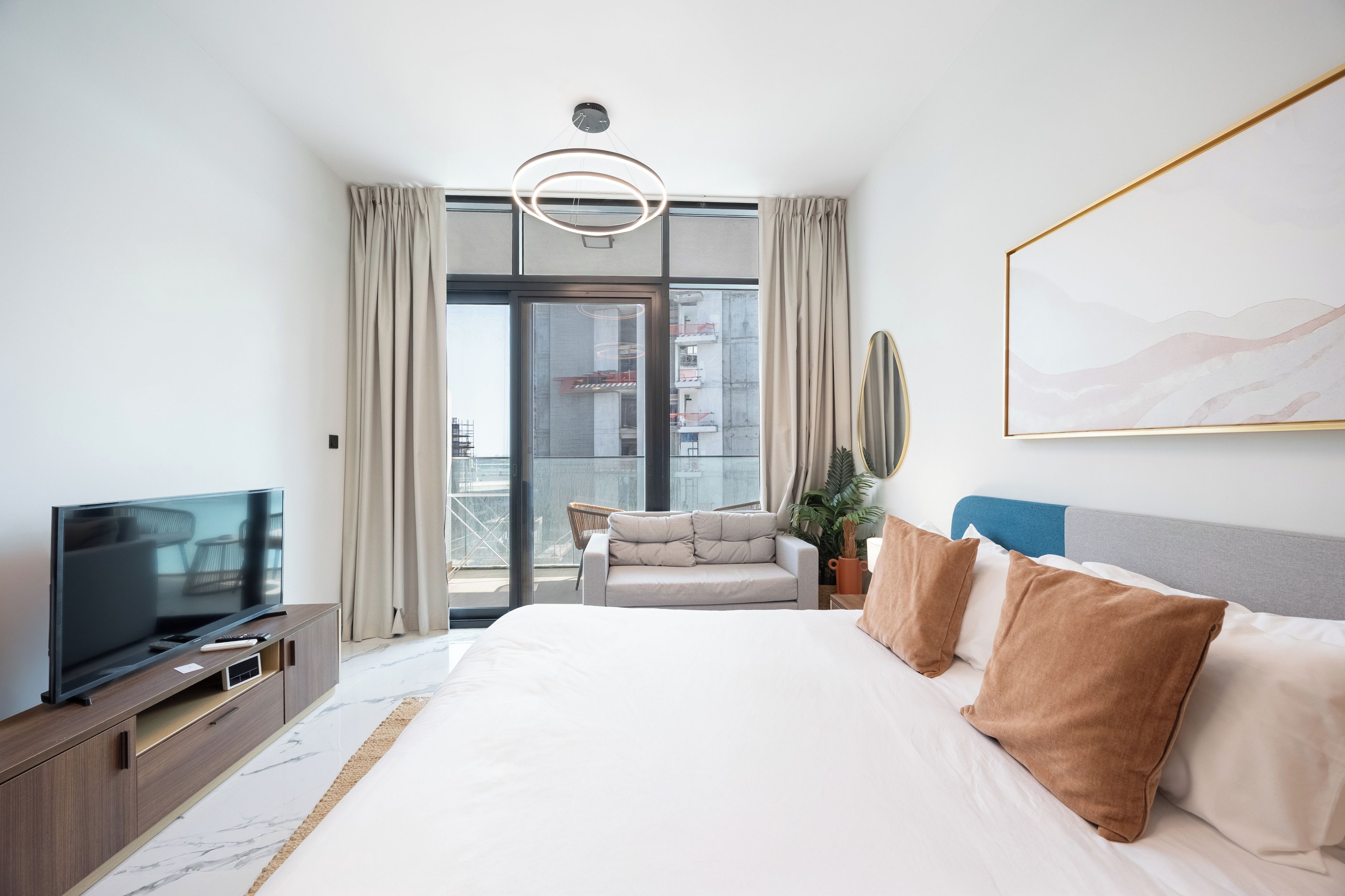 A modern hotel room with a large bed, a television, a sofa, and a balcony with a view of buildings.