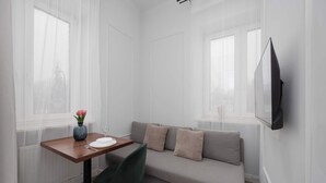 A modern living room with a sofa, a dining table with a vase of flowers, and a flat-screen TV mounted on the wall. - Cozy Studio for Two | Close to Metro | Pet-Friendly (Warszawa)