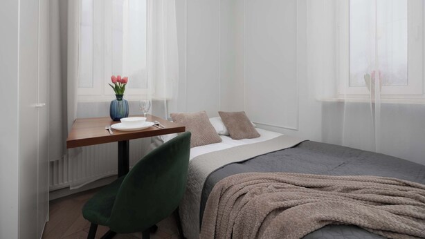 A bedroom with a bed, a bedside table with a vase of flowers, a chair, and a window with sheer curtains. - Cozy Studio for Two | Close to Metro | Pet-Friendly (Warszawa)