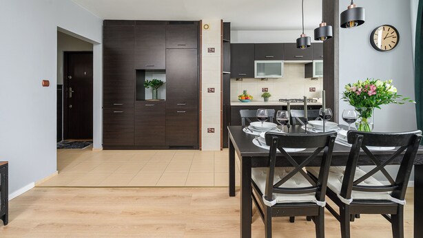 A modern kitchen with dark cabinets, a dining area with a wooden table and chairs, and a clock on the wall. - Lumina 3-rooms apartment with parking (Warsaw)