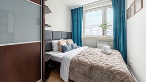 A neatly arranged bedroom with a bed, a window with curtains, a radiator, and a potted plant. - Lumina 3-rooms apartment with parking (Warsaw)
