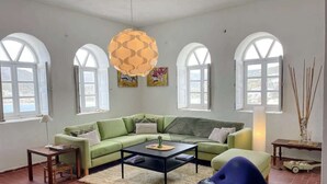 Un salon avec un canapé vert, une table basse noire et un fauteuil bleu. Il y a des fenêtres avec des cadres blancs et un luminaire suspendu.