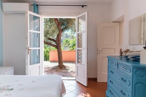 Eine geöffnete Tür, die auf einen Balkon mit einem Baum führt, ein Bett mit floralem Überwurf und eine blaue Kommode mit Schubladen.