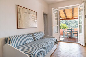 Ein Zimmer mit einem gestreiften Sofa, einem gerahmten Bild an der Wand und einer offenen Tür, die auf einen Balkon mit Holzboden und Blick auf Grün führt.