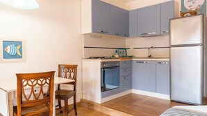 A compact kitchen with grey cabinets, a white refrigerator, a stove, and a dining area with a wooden chair and table. - Sea walking distance, cozy apartment with garden, pets welcome (Pomonte)