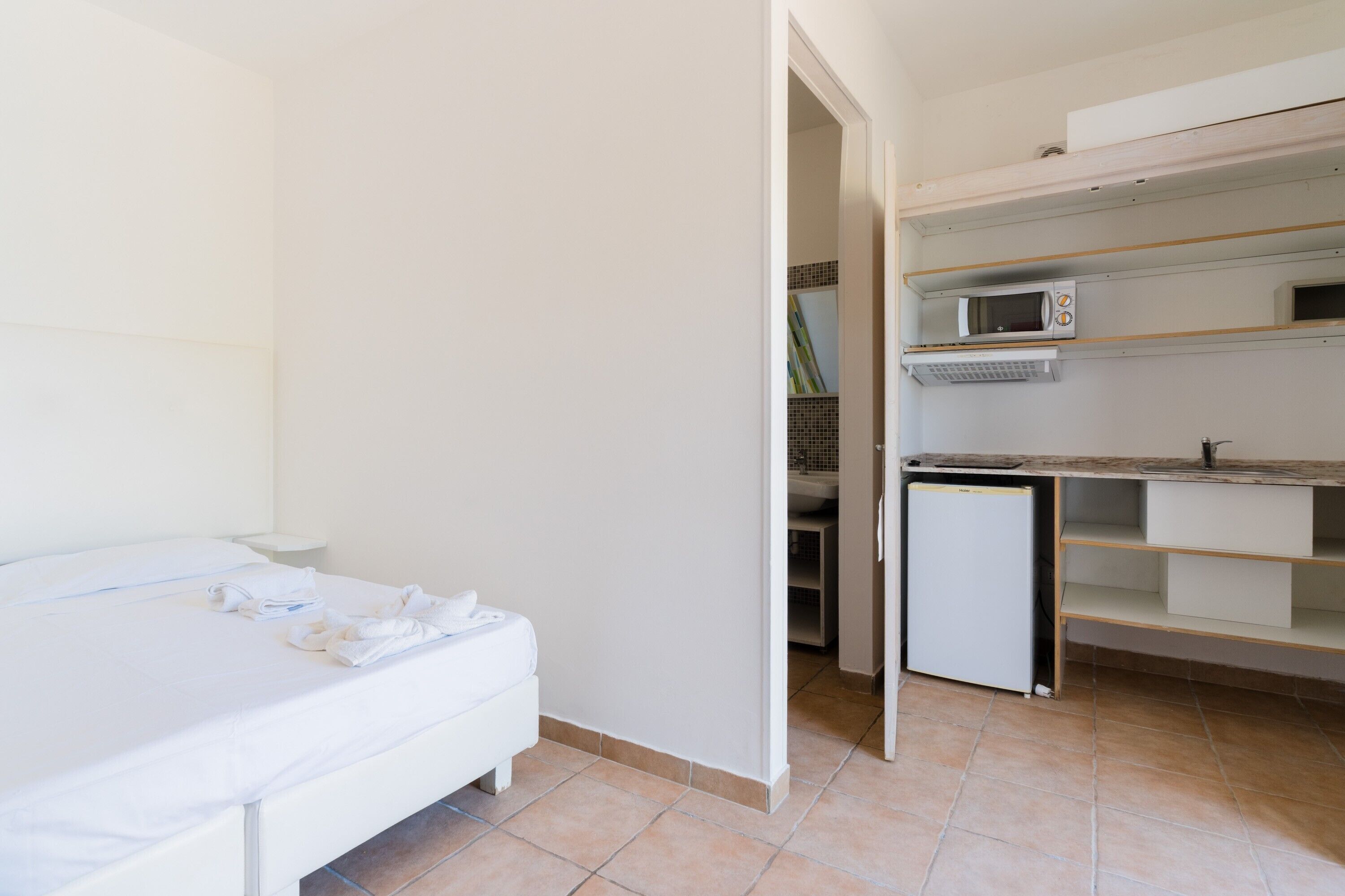 A bedroom with a bed, a small kitchenette with a microwave and a refrigerator, and a bathroom visible through an open door.