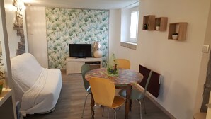 Un salon chaleureux avec un canapé, une table à manger, des chaises, un téléviseur et un mur orné d’un motif végétal.