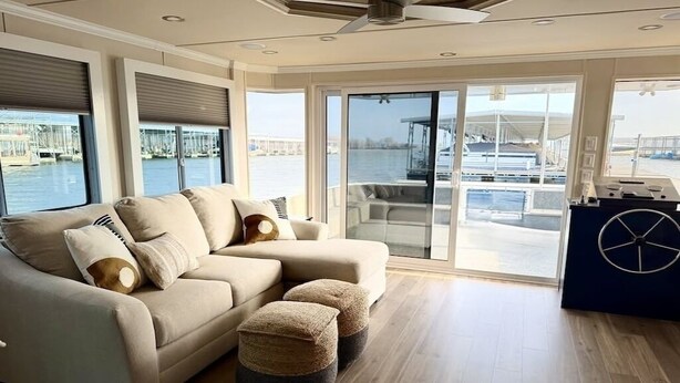 A modern living room with a sofa and ottomans, large windows with a marina view, and a ceiling fan. - 4-bedroom houseboat in historic Old Kuttawa, KY (Kuttawa)