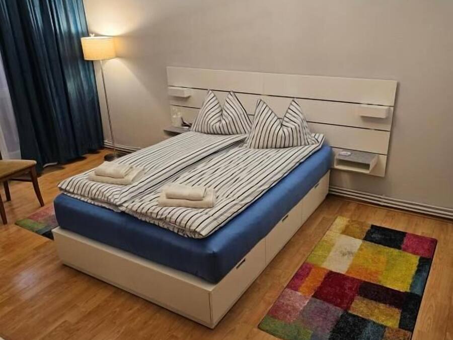 Una cama bien hecha con ropa de cama a rayas y almohadas, una mesita de noche, una lámpara y una alfombra colorida sobre un piso de madera.