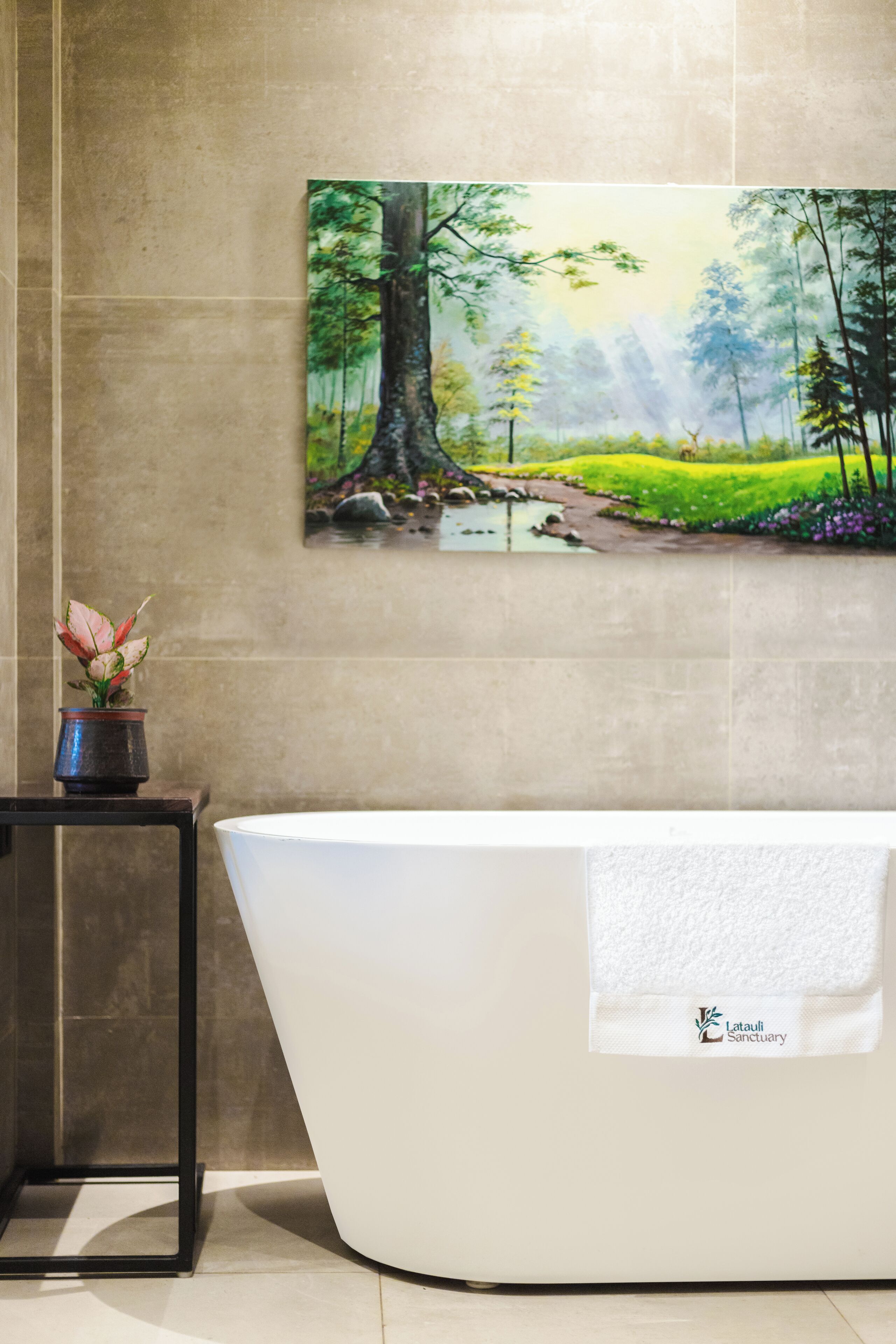 Un baño moderno con una bañera independiente, un cuadro de una escena boscosa y una mesita con una planta en maceta.