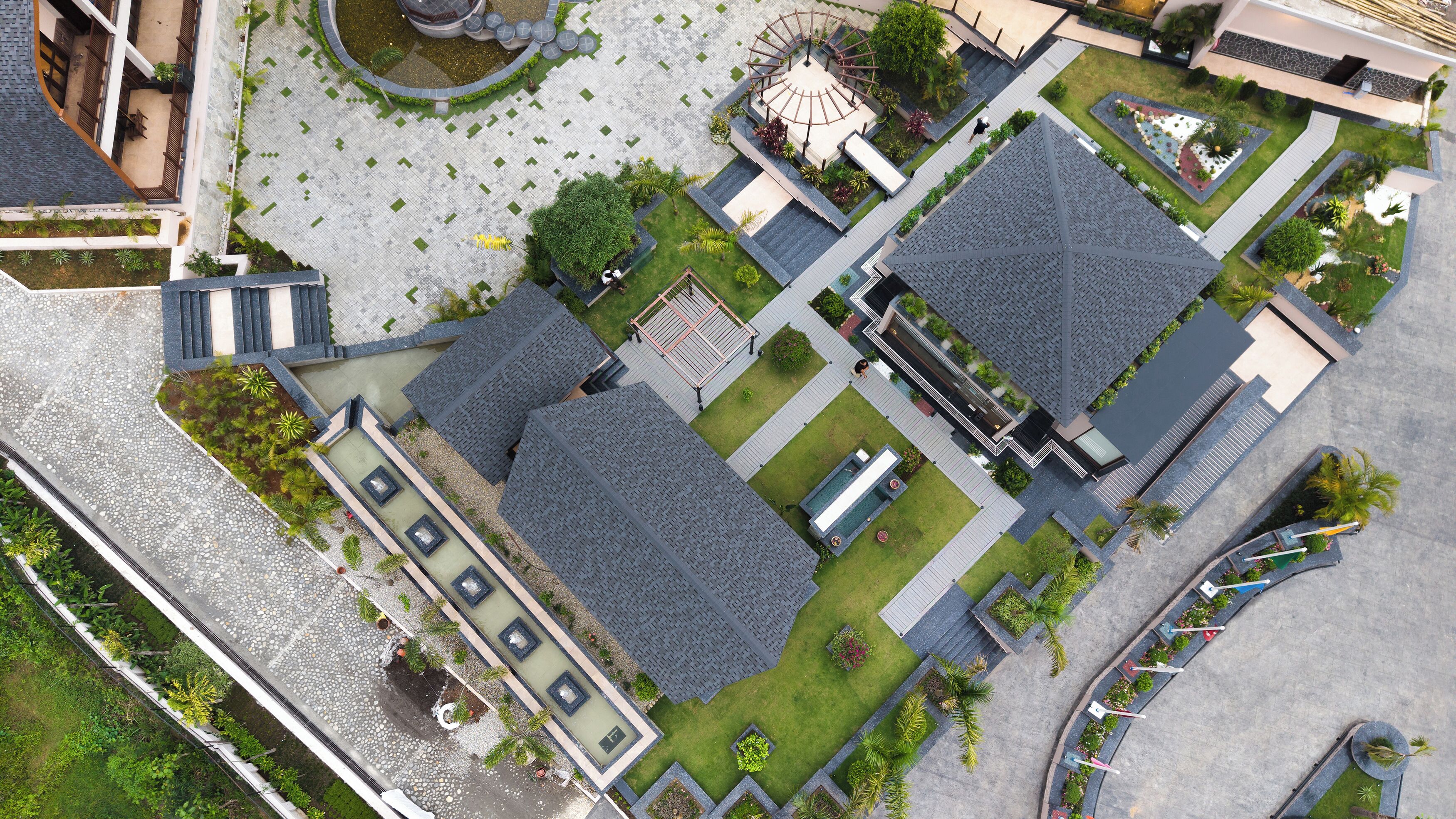 An aerial view of a modern building complex with multiple roofed areas, landscaped gardens, and a central circular structure.