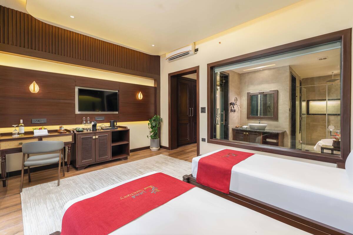 Habitación de hotel con dos camas, televisión, escritorio con silla, un espejo grande y baño visible a través de una puerta abierta.