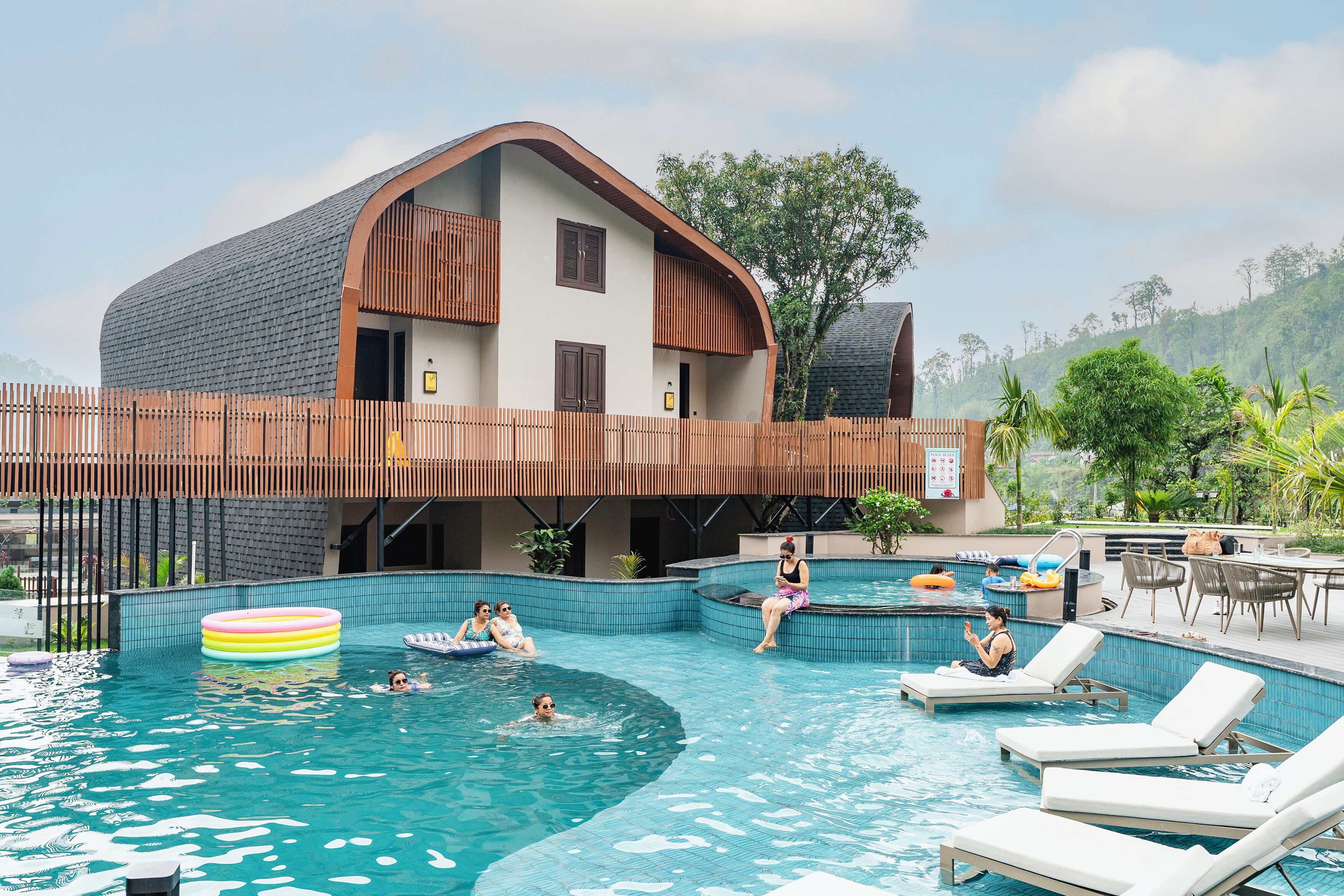 A modern building with a curved roof, a swimming pool with lounge chairs, and a wooden deck.
