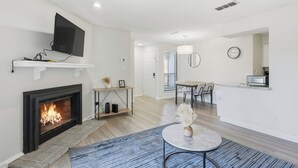Ein modernes Wohnzimmer mit Kamin, einem an einer Wandregal montierten Flachbildfernseher, einem Essbereich mit Tisch und Stühlen sowie einer Küchenzeile mit Mikrowelle.