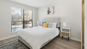 Ein Schlafzimmer mit einem großen Bett, einem Nachttisch mit Lampe, einem Fenster mit Jalousien und einem kleinen Wandbild.