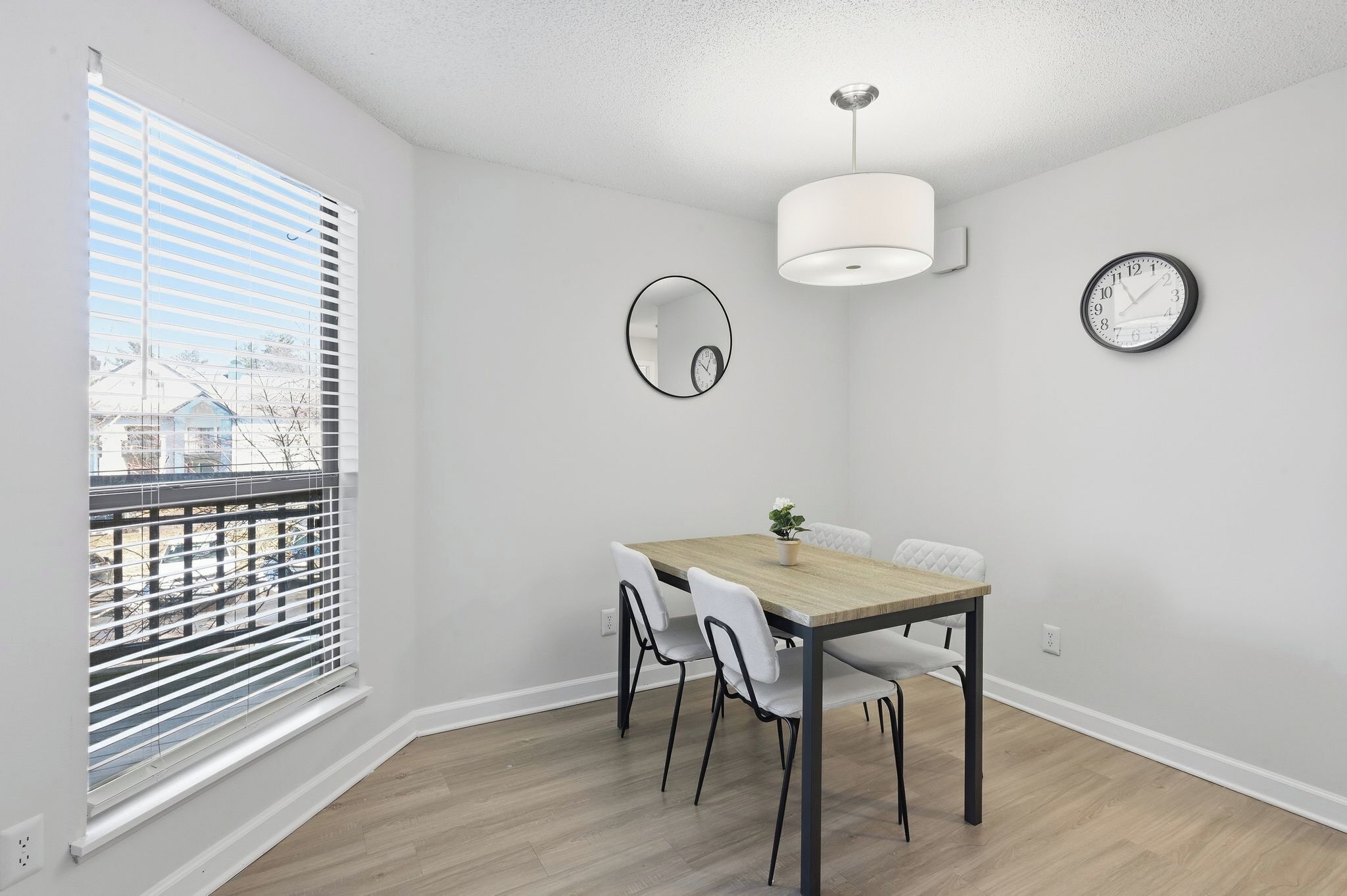 A dining area with a wooden table, white chairs, a round mirror, a wall clock, and a ceiling light fixture.