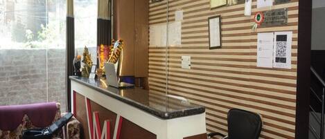 A hotel reception area with a reception desk, a clock, and various posted notices including no smoking and hand wash signs.