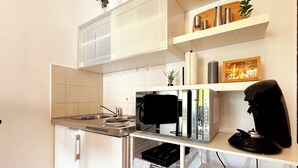 Una cucina compatta con forno a incasso, microonde e macchina del caffè. Sopra il bancone, mensole con stoviglie e una piccola pianta.